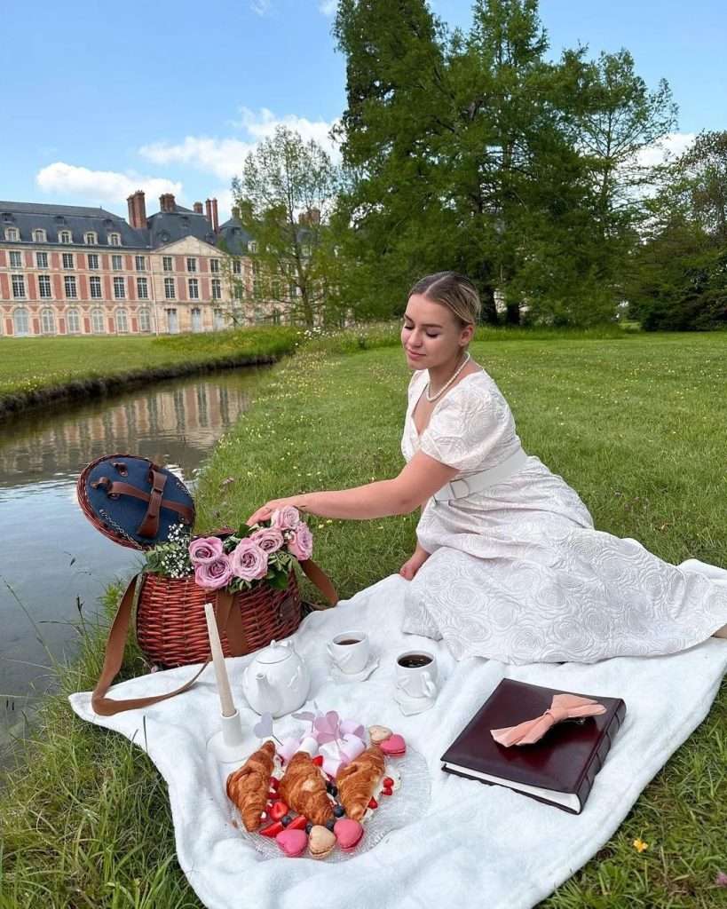 Solo picnic in a peaceful park with a blanket, basket of fruits, and a book under a clear sky.